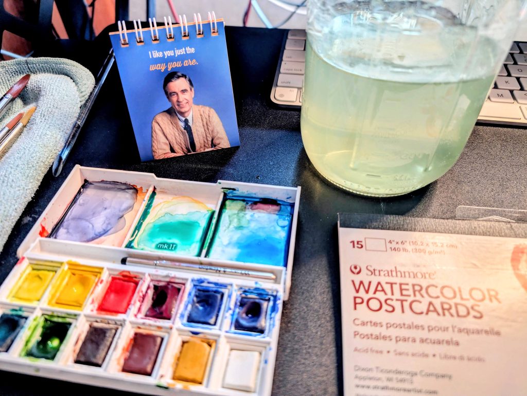 Watercolor paint set with various colors, paintbrushes, watercolor postcards, and a jar of water on a desk.